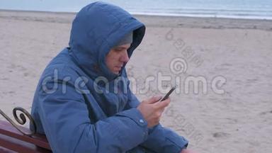 穿着蓝羽绒服坐在沙滩上的长凳上，在手机上的社交媒体上写一篇文章的人博主